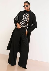 Must have villaneeritud must villane mantel leopardimustrilise kampsuni, laiade mustade pükste ja mustade prillide kohal. Omab sileda tekstuuri ja täpset lõiget.