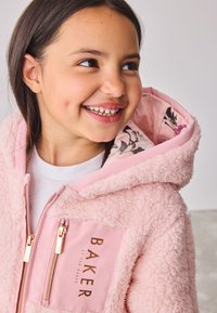 Smilende pige iført en pink fluffy jakke med blomsterfor i hætten og gyldne lynlåsfunktioner, der kigger til siden.
