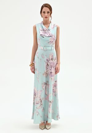 Femme portant une robe longue fleurie sans manches, ceinturée, aux tons pastel bleu et rose, debout les mains détendues le long du corps sur un fond blanc.