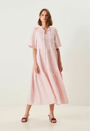 Nainen seisoo yksivärisen taustan edessä, hänen yllään on vaaleanpunavalkoraidallinen midimekko, jossa on liehuvat hihat, ja jalassaan punaiset korolliset sandaalit.