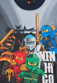 Szürke póló, amelyen élénk színekben láthatóak a LEGO Ninjago karakterei: fekete, piros, zöld, kék és fehér, kardokkal, lángokkal és stilizált háttérrel.