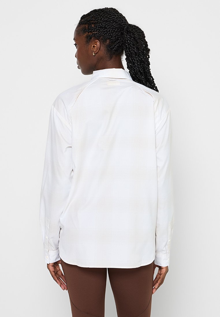 Biała koszula z długim rękawem o subtelnym wzorze w kratkę, z mankietem zapinanym na guziki i podszewką z tyłu. Gładka tkanina, luźny krój.