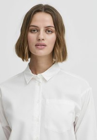 Chemise blanche à boutons avec un col et une poche poitrine simple. Fabriquée en tissu lisse, avec des manches longues et une coupe ajustée.