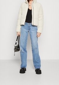Nafoukaná bunda v off-white barvě, černý top, světle modré džíny rovného střihu a robustní černé platformové boty, držící malou černou kabelku.