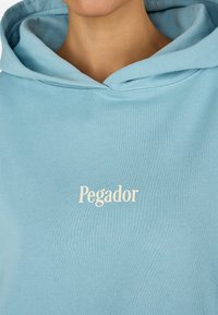 Ανοιχτό μπλε φούτερ με απαλή υφή, ρυθμιζόμενη κουκούλα με κορδόνι και λογότυπο "Pegador" σε κρεμ ράψιμο.