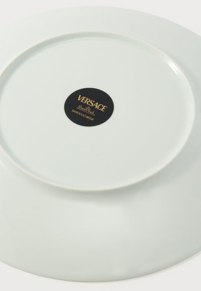 Λευκό κεραμικό πιάτο αναποδογυρισμένο, με μια μαύρη κυκλική ετικέτα Versace με χρυσό κείμενο στο κέντρο.