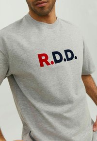 Sivá bavlnená tričko s krátkymi rukávmi, s výraznými červenými a námorníckymi písmenami "R.D.D." na hrudi. Mäkka textura, pohodlný strih.