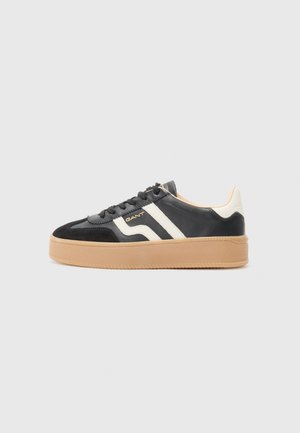 Black leather sneakers with white stripes and a gum rubber sole. Features rounded toe and lace closure with a textured upper. Branding displayed.