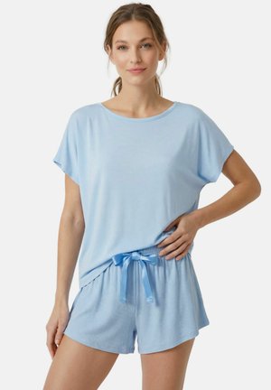 Femme en haut bleu clair à manches courtes et short assorti avec un ruban, debout avec une main sur la hanche, fond neutre.