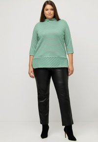 Grün-weiß gestreiftes Langarmoberteil mit hohem Kragen und asymmetrischem Saum, kombiniert mit schwarzen Kunstlederhosen und schwarzen Stiefeletten.