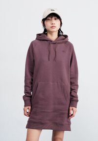 Pullover hettekjole i dempet lilla bomullsblanding, med en frontlomme, snorhette og ribbestrikket kant. Modellen har på seg en beige caps.