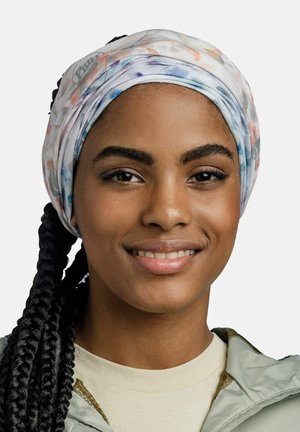Smiling woman with braided hair wearing a pastel-colored headscarf and light-colored jacket on a plain background.