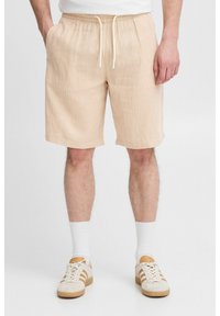 Beige shorts dengan pola bergaris yang bertekstur, dilengkapi dengan pinggang elastis dan tali serut. Dipasangkan dengan kaus kaki putih dan sepatu kets beige.