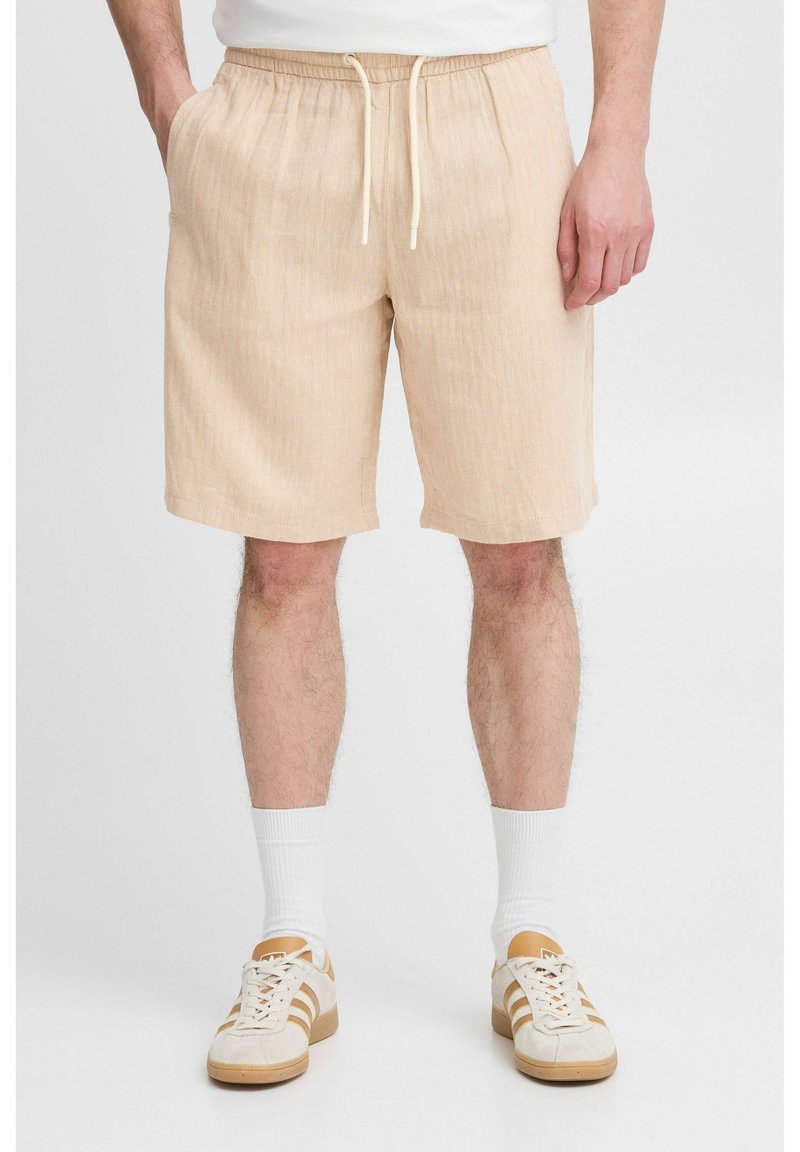 Beige shorts dengan pola bergaris yang bertekstur, dilengkapi dengan pinggang elastis dan tali serut. Dipasangkan dengan kaus kaki putih dan sepatu kets beige.