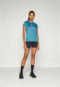 Turkusowa koszulka z krótkim rękawem odprowadzająca wilgoć z grafiką gór; w komplecie z czarnymi szortami i skarpetkami sięgającymi kostek. Czarne buty sportowe.