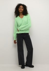 Mintgroene oversized trui met een V-hals en ribgebreide manchetten, gecombineerd met hoge zwarte jeans en zwarte schoenen.