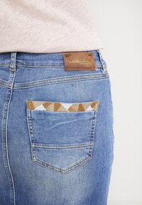 Blå denimkjol med en brun läderlapp märkt "Mos Mosh" och en unik mönstrad tygdetalj som syns från bakfickan.