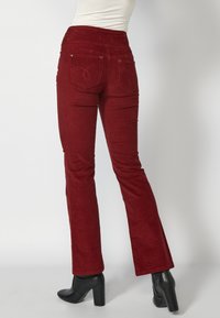 Vue arrière d'une personne portant un pantalon flare en velours côtelé rouge foncé taille haute et des bottines noires à talons sur fond blanc.