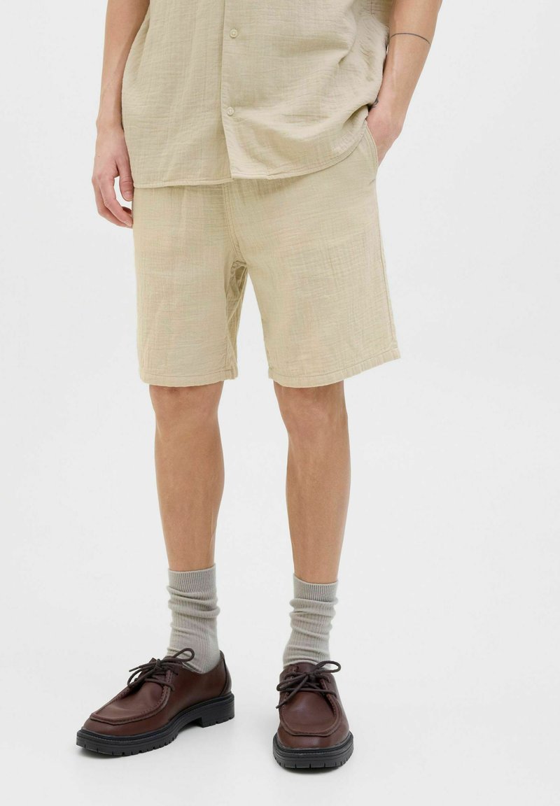 Pantalones cortos beige con textura y un ajuste relajado, combinados con zapatos marrones y calcetines gris claro. La tela tiene un aspecto suave y ligeramente arrugado.