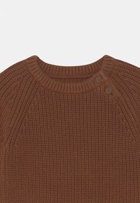 Brun stickad tröja med ett texturerat ribbmönster, rund halsringning och två bruna tryckknappar på vänster axel.