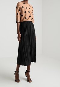 Kvinna iklädd en beige långärmad topp med svarta prickar, en svart plisserad midikjol och svarta högklackade ankelremssandaler.