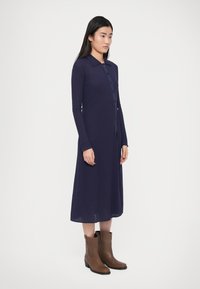 Tmavomodré rebrované šaty s dlhými rukávmi, špicatým golierom, prednými gombíkmi a celodĺžkovým strihom, skombinované s hnedými členkovými čižmami.