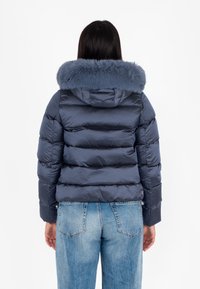 Giacca blu navy voluminosa con cappuccio in pelliccia morbida, azzurro chiaro. Presenta sezioni trapuntate orizzontali e chiusura con zip. Indossata sopra jeans azzurri chiari.
