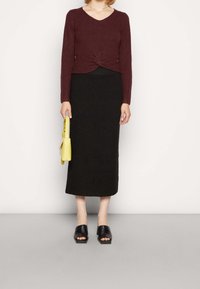 Borduren geribbelde langmouwen top in maroon met een gedraaide voorkant, zwarte gebreide midi rok, zwarte instapsandalen en een gele handtas.