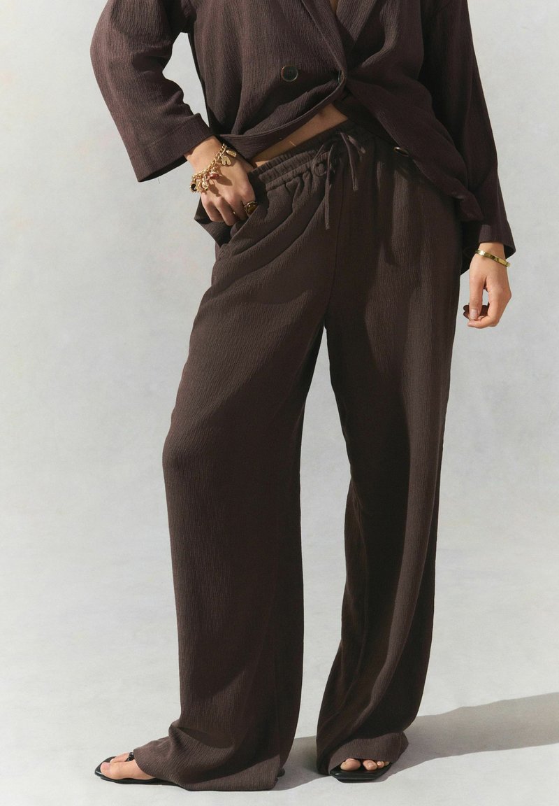 Brown, textured wide-leg pants with a drawstring waistband, paired with a matching blazer. Accessorized with gold jewelry and open-toe sandals.