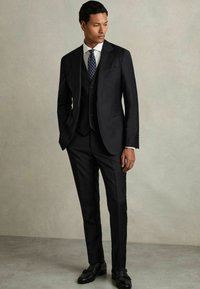 Costum negru în trei piese, cu sacou și vestă adaptate. Cămașă albă și cravată bleumarin cu puncte. Complementat de pantofi negri lucioși.