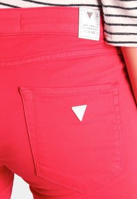 Pantalones de mezclilla rojos con un bolsillo trasero y una etiqueta de logo en forma de triángulo plateado. El material tiene una textura suave con detalles de costura mínimos.
