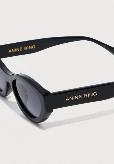 Crne ovalne sunčane naočale s tamnim lećama na kojima je zlatnim slovima ispisano "ANINE BING" na oba kraka.