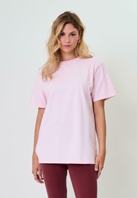 Světle růžové bavlněné tričko, kulatý výstřih, volný střih, krátké rukávy, hladká textura, v kombinaci s burgundskými kalhotami. Žádné viditelné vzory ani akcenty.