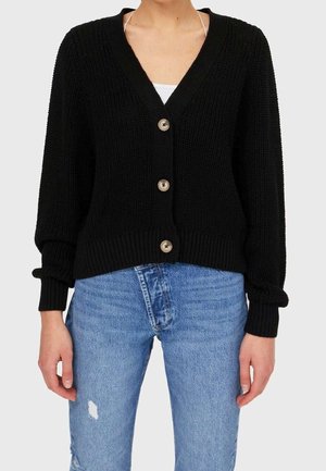 Personne portant un cardigan en maille noire avec trois boutons sur un haut blanc et un jean bleu usé sur un fond neutre.