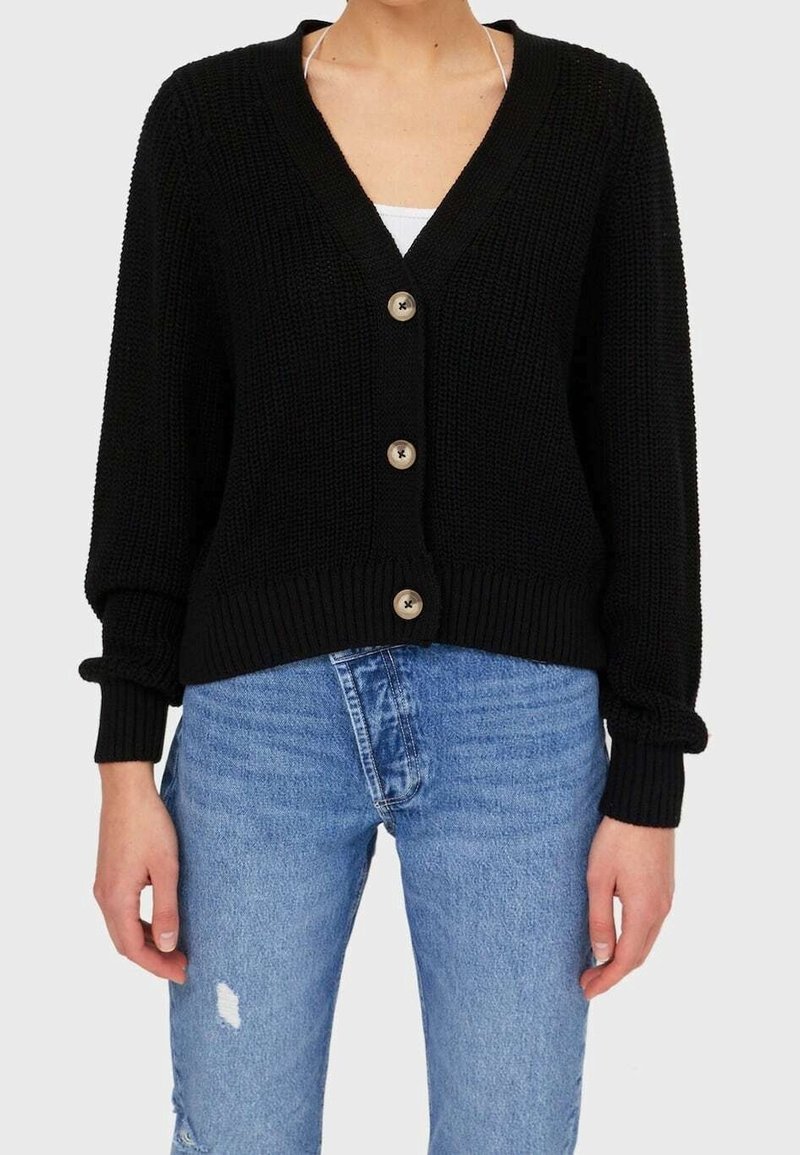 Personne portant un cardigan en maille noire avec trois boutons sur un haut blanc et un jean bleu usé sur un fond neutre.
