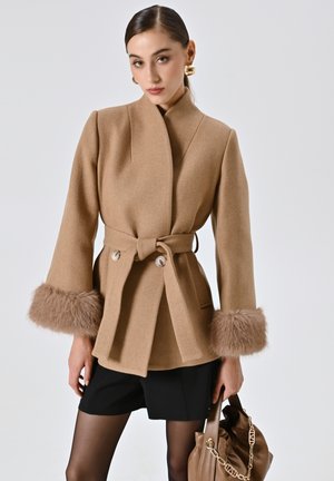 Beige Wollmantel mit Wickeldesign, Seilbindegurt und Pelzaufschlägen. Verfügt über große Knöpfe und eine glatte Textur. Kombiniert mit schwarzen Shorts und einer braunen Handtasche.
