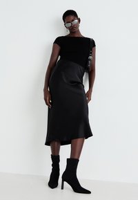 Top negro de manga corta combinado con un vestido midi de satén negro. La modelo lleva botas de tobillo negras y unas gafas de sol elegantes, con un diseño minimalista.