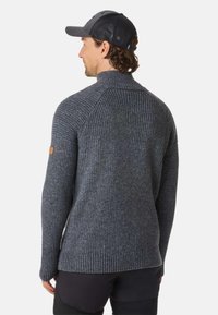 Pull en maille gris avec col haut et manches raglan, arborant un patch en cuir sur le bras gauche. Porté avec un pantalon noir.