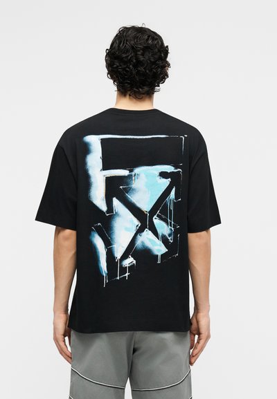 Άτομο με σγουρά μαλλιά που φοράει μαύρο μπλουζάκι με μεγάλο αφηρημένο μπλε και λευκό γκράφιτι στο πίσω μέρος, συνδυασμένο με γκρι σορτς.