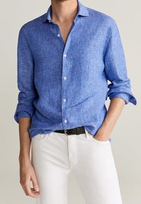 Camisa azul de lino con botones, acabado texturizado, mangas largas remangadas y pantalones blancos. Cuenta con un cuello clásico y botones al frente.