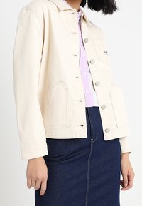 Kvinna klädd i en krämfärgad knappjacka över en lavendelfärgad skjorta och en mörkblå denimkjol mot en enhetlig bakgrund.