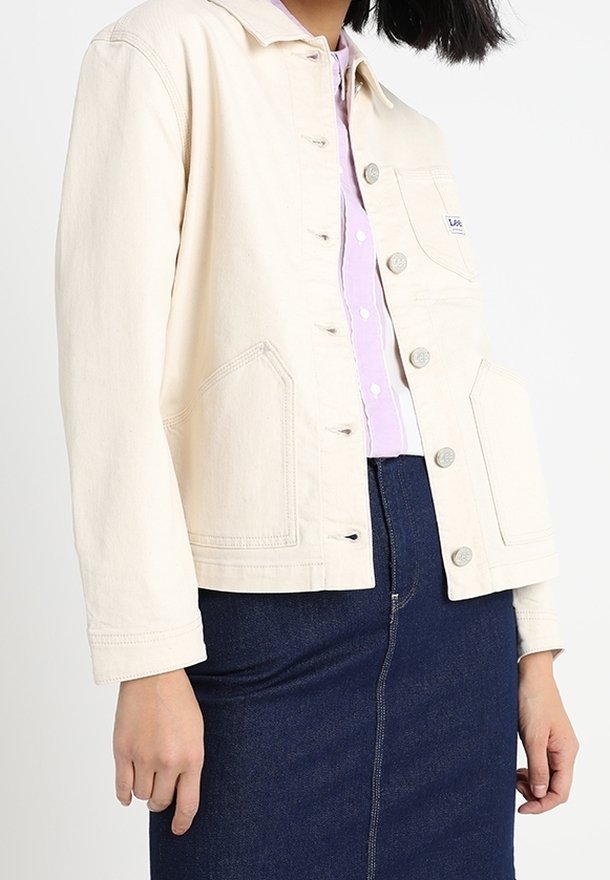 Kvinna klädd i en krämfärgad knappjacka över en lavendelfärgad skjorta och en mörkblå denimkjol mot en enhetlig bakgrund.