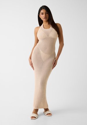 Mujer de pie con un vestido maxi de punto acanalado beige sin mangas y sandalias blancas tipo slide sobre un fondo claro y liso.