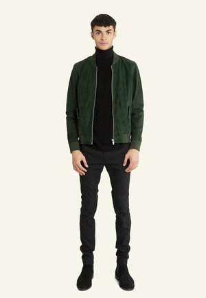 Veste bomber en daim vert, fermeture éclair à l'avant, poignets côtelés, portée sur un col roulé noir, associée à un pantalon noir slim et des bottines noires.