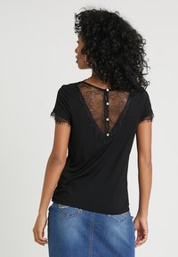 Haut noir à manches courtes avec un dos en dentelle, présentant un décolleté en V et des boutons décoratifs le long du centre. Les bordures en dentelle sur les manches ajoutent une touche de détail.