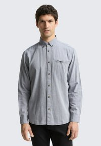 Chemise à carreaux bleu clair avec manches longues, col boutonné, poche poitrine et boutons marron. Associée à un pantalon noir.