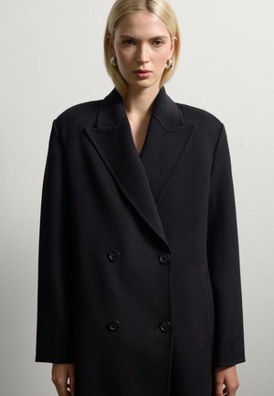 Woman with straight blonde hair wears oversized black double-breasted coat and silver hoop earrings against plain light background.