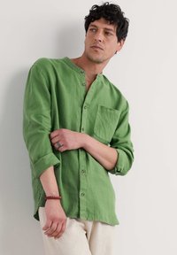 Camicia di lino verde con colletto alla mandarina, abbottonatura frontale e taschino sul petto. Le maniche lunghe sono arrotolate, mostrando una consistenza morbida.