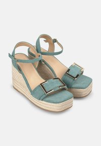 Une paire de sandales à talons compensés en suede bleu-vert, avec une large bride ornée d'un accent métallique, une semelle en jute tressé et des brides de cheville réglables.