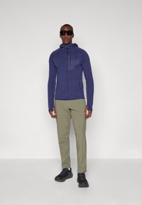 Veste de sport zippée navy avec un panneau lisse sur la poitrine, associée à un pantalon beige et des baskets noires. Le mannequin porte des lunettes de soleil noires.